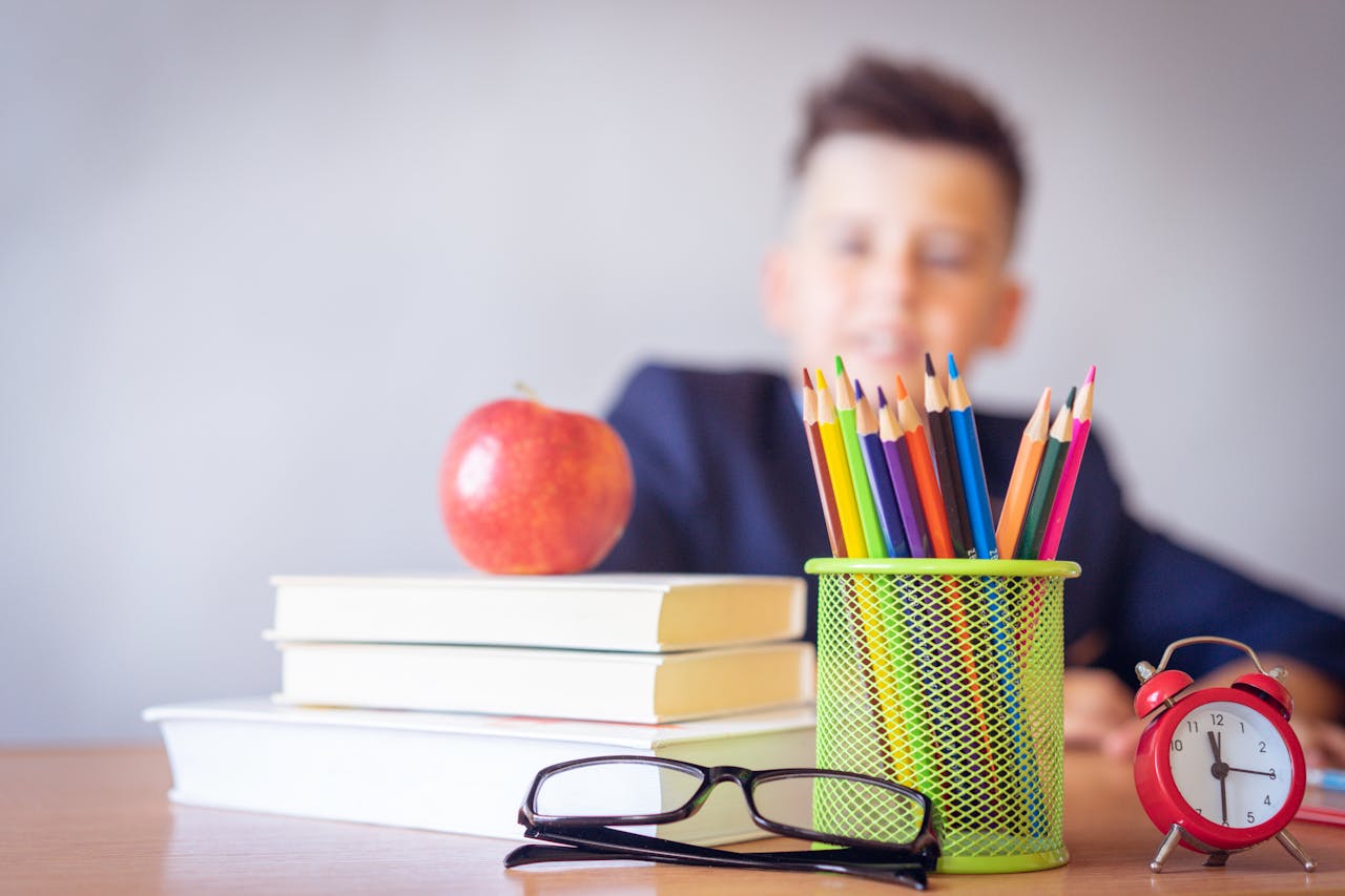 Schoolboy smiling behind a desk with books, pencils, and an alarm clock symbolizing study and creativity.